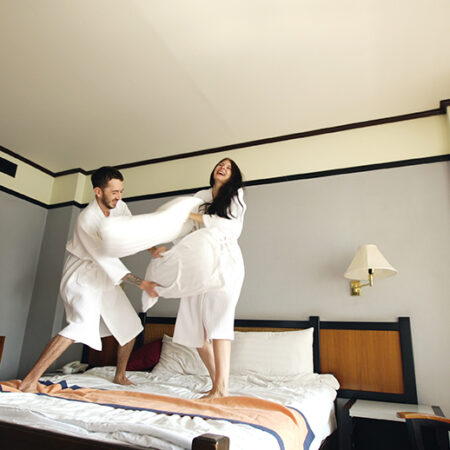 Young happy couple in bathrobe fight pillows on bed in hotel during their honeymoon